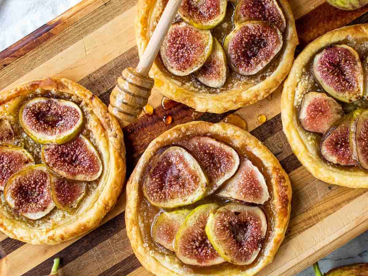 puff pastry fig tartlets on a wooden board with honey dipper drizzling honey over pastries viewed from above.