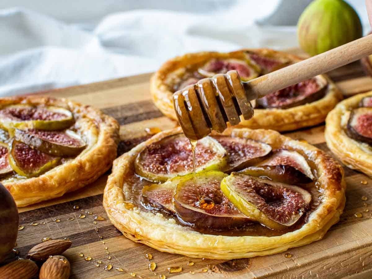 puff pastry fig tartlets on a wooden board with honey dipper drizzling honey over pastries.