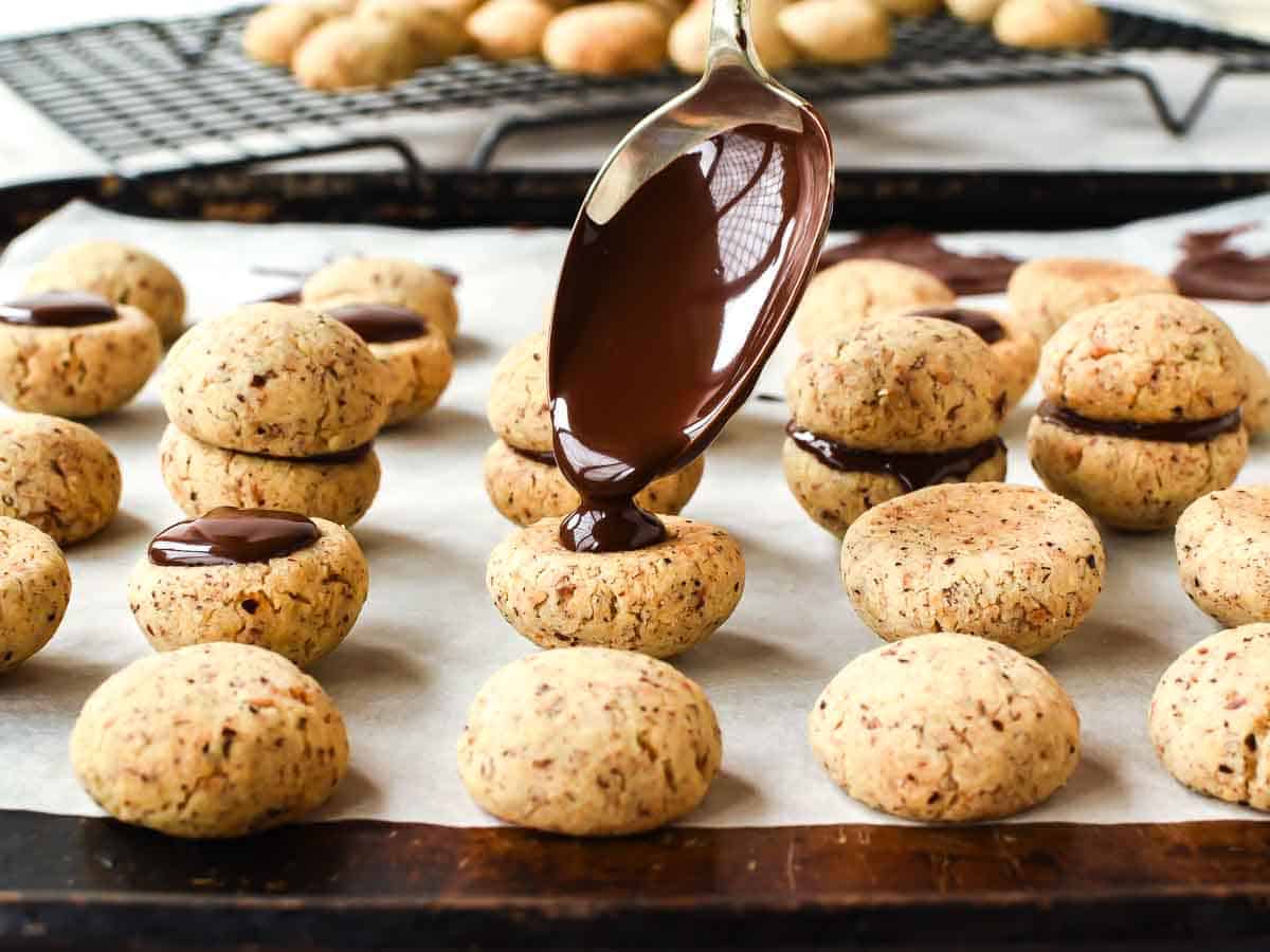 hazelnut cookies on a baking tray with a spoon pouring a little chocolate on a cookie.