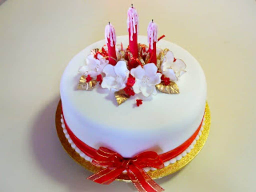 cake covered with white fondant icing topped with red candles, white flowers and gold leaves wrapped with red ribbon.