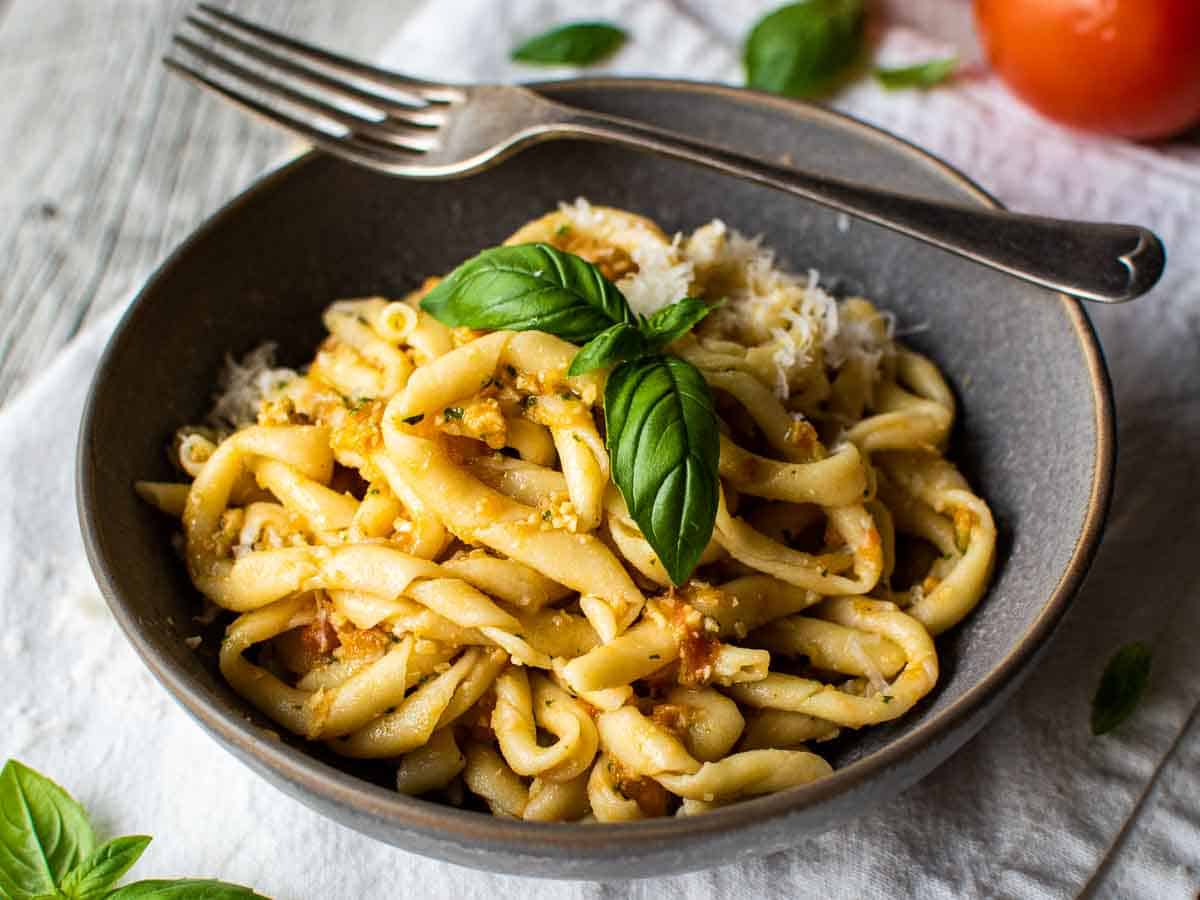 A grey bowl filled with handmade, twirled pasta with a reddish sauce, topped with a sprig of basil and a fork balance on the edge of the bowl.