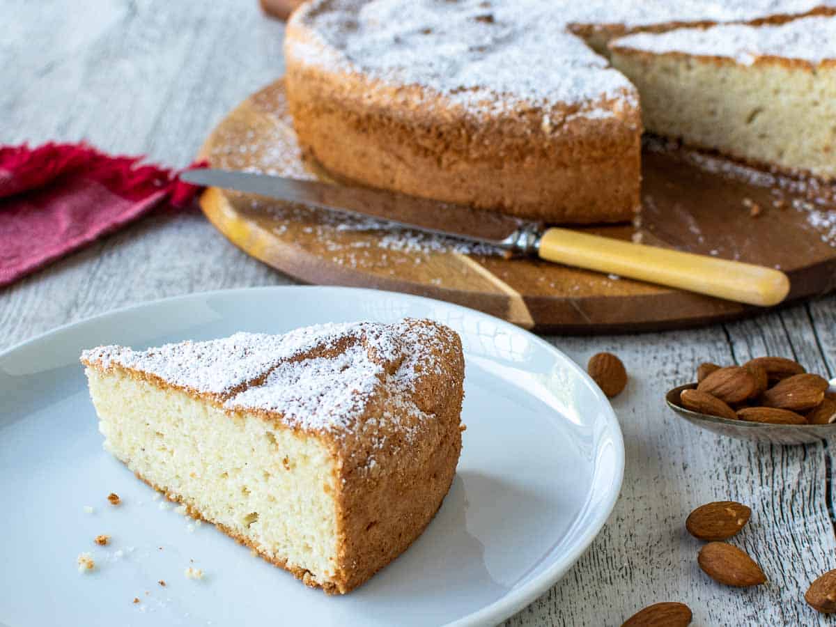 Wedge of cake on a white plate with whole cake in the background.