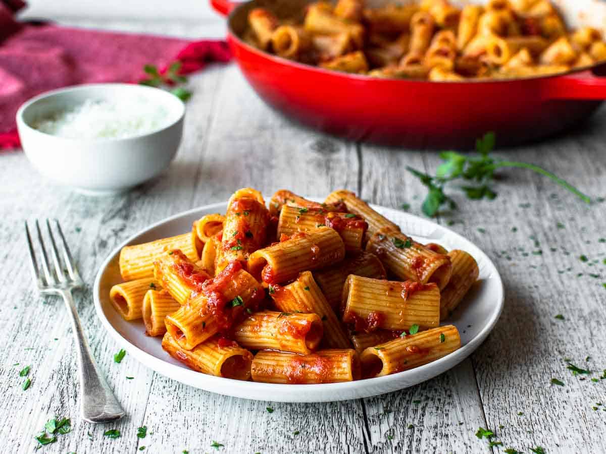 Rigatoni with tomato pasta sauce on a white plate with fork and red skillet in the background.