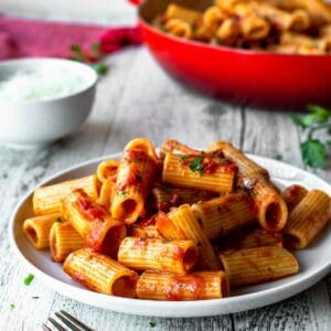 Rigatoni Arrabbiata on a white plate with red skillet in the background.