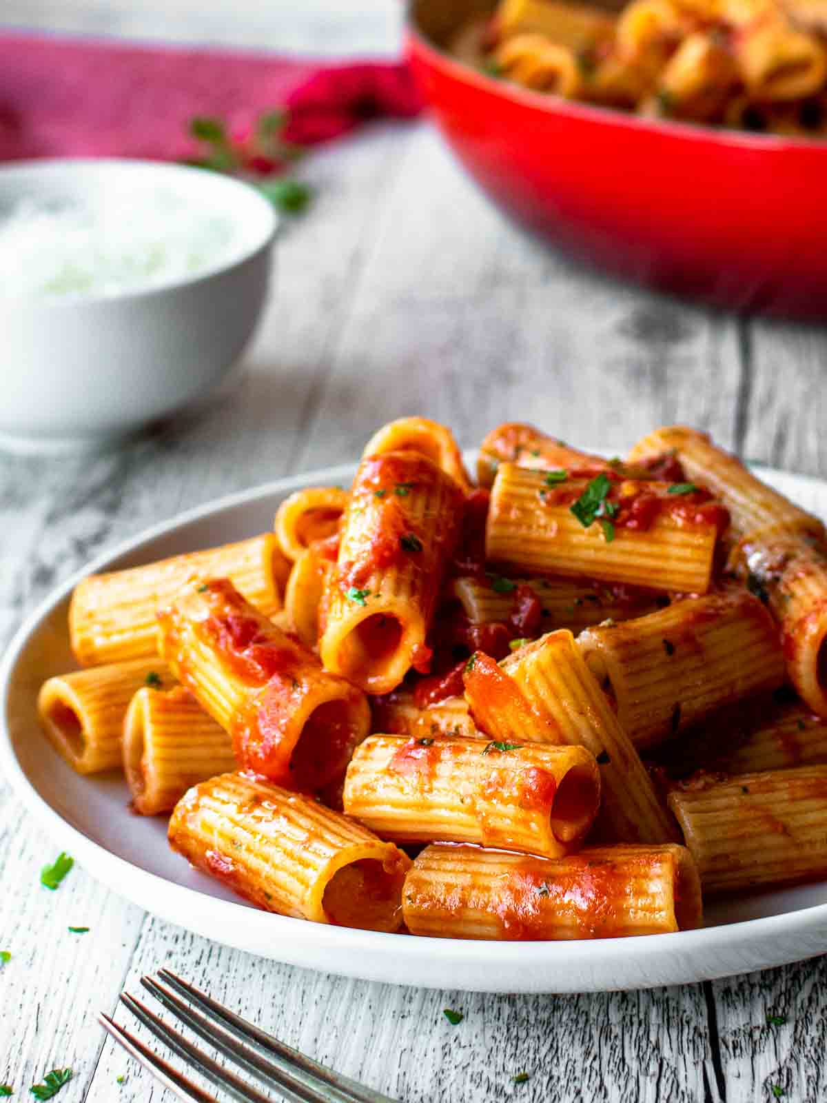 Rigatoni Arrabbiata on a white plate with red skillet in the background.