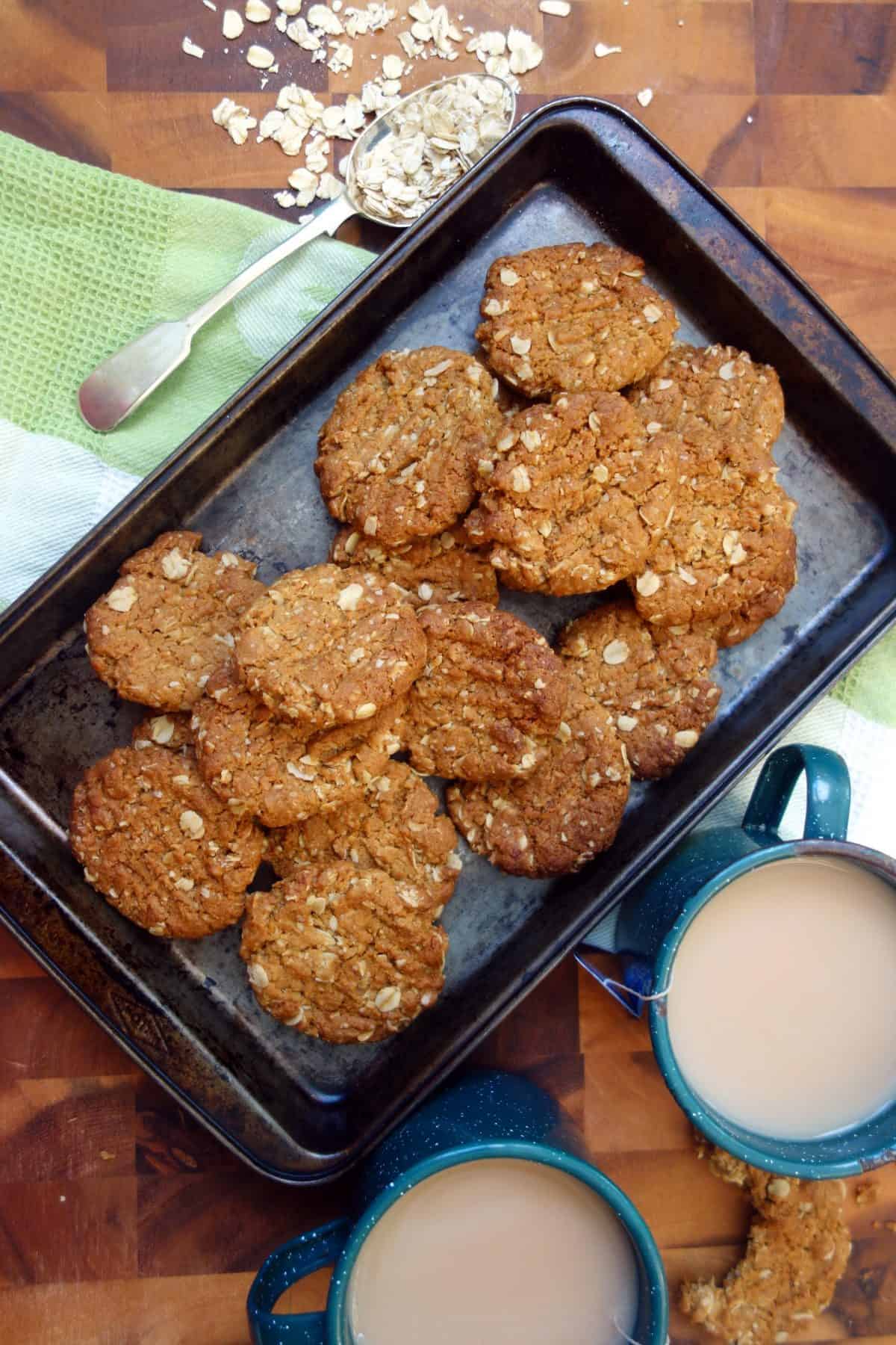 These Anzac Biscuits are beautifully flavored with golden syrup and ground ginger. #anzacbiscuits #cookies #biscuits #recipe #australianrecipe Overhead view of Anzac Biscuits in dark coloured baking tray