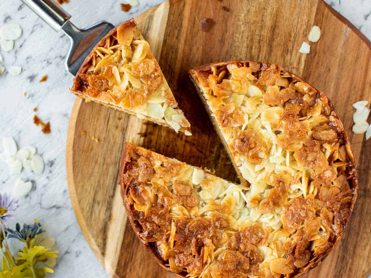 slice of cake with almond topping cut from whole cake on a wooden board viewed from above.