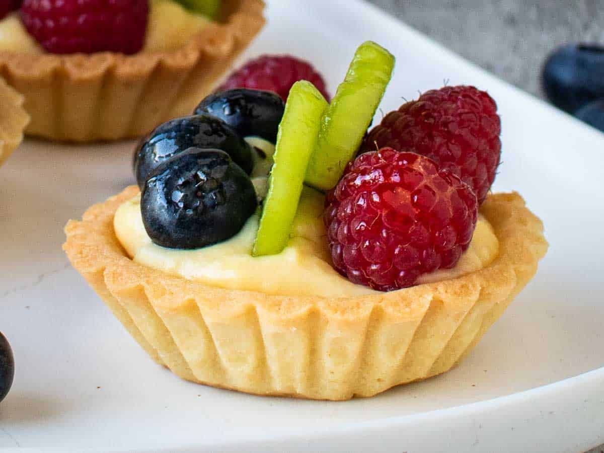 Close up of fluted tart filled with pastry cream and topped with blueberries, sliced kiwi fruit and raspberries