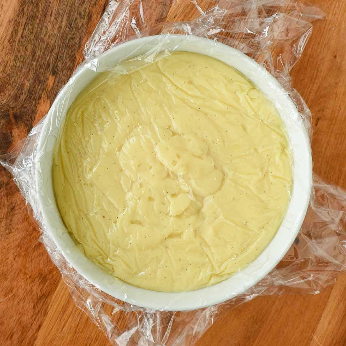 Thickened pastry cream in a white bowl covered with plastic wrap.