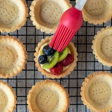 Individual fruit tarts filled and topped with fruit being brushed with a red pastry brush.