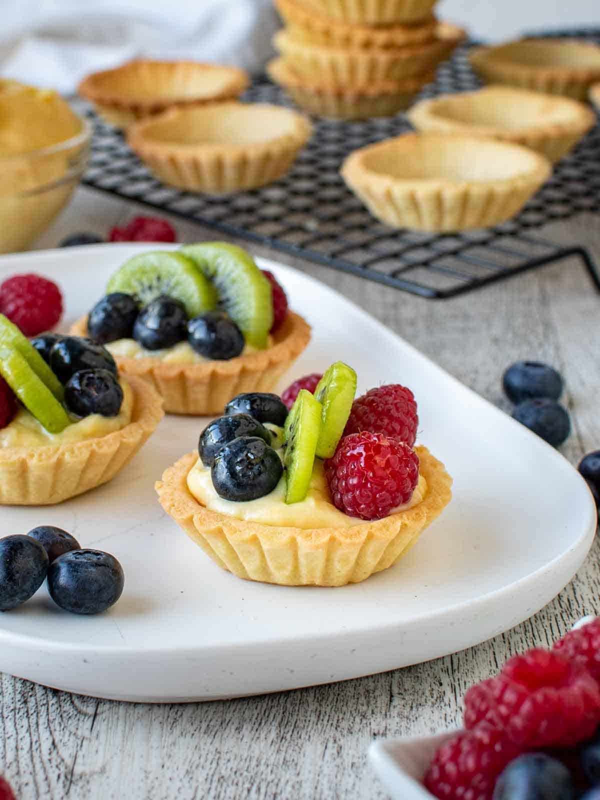 Mini fruit tarts on a white oblong plate with tartlet bases in the background.