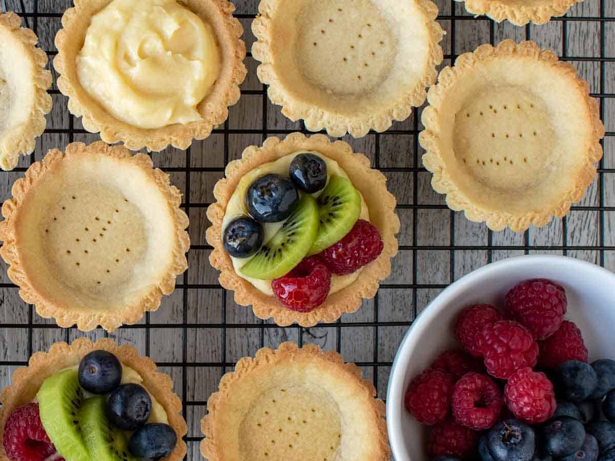 Overhead view of mini tarts, some empty, some filled with pastry cream and some topped with berries and sliced kiwi fruit.