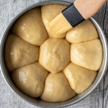 Nine balls of dough well risen in a cake pan being brush with egg.