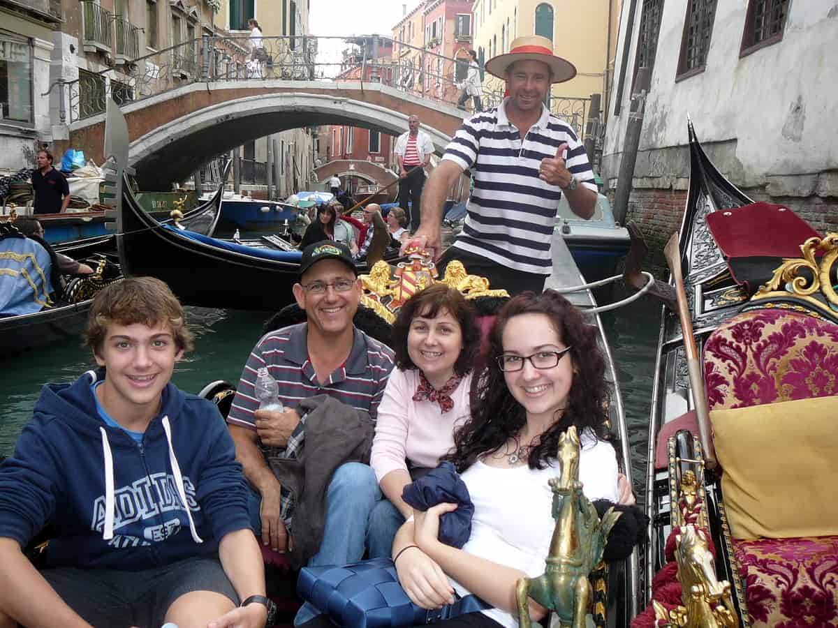 boy, man, woman and a girl in a Venetian gondola with man in black and white striped shirt in the background. 