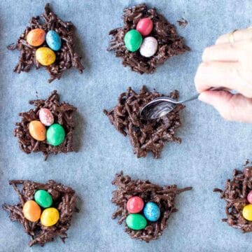 Chocolate noodle nests with candy eggs in the middle on a baking sheet with one nest being shaped with a spoon viewed from above.