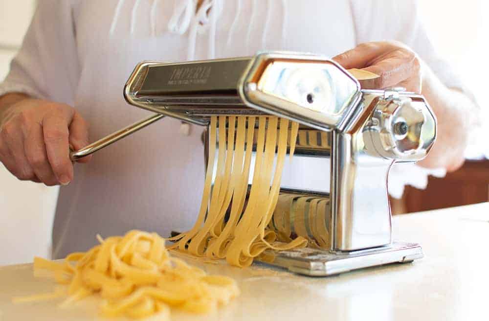 woman in white using a pasta machine to cut pasta sheets into long strips.