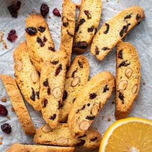 Orange Cranberry Biscotti on white paper with halved oranges and dried cranberries scattered viewed from above.