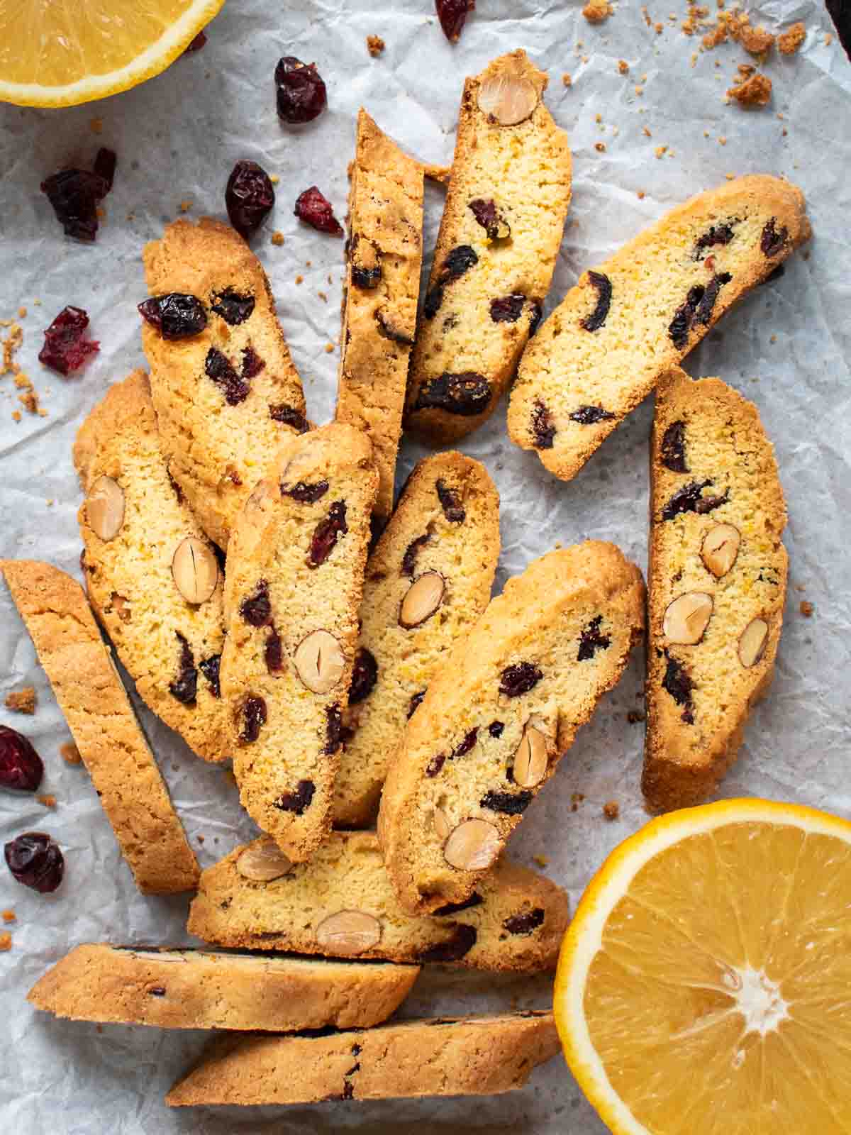 Orange Cranberry Biscotti on white paper with halved oranges and dried cranberries scattered viewed from above.