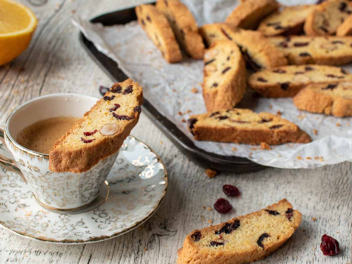 Biscotti on a paper covered baking sheet and one perched on the edge of a coffee cup.