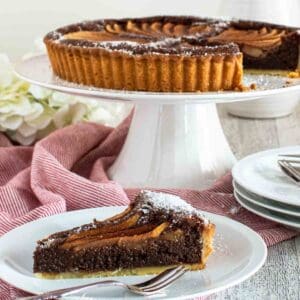 Chocolate almond pear tart on a white cake stand with a slice removed and plated in front.