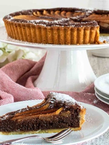 Chocolate almond pear tart on a white cake stand with a slice removed and plated in front.