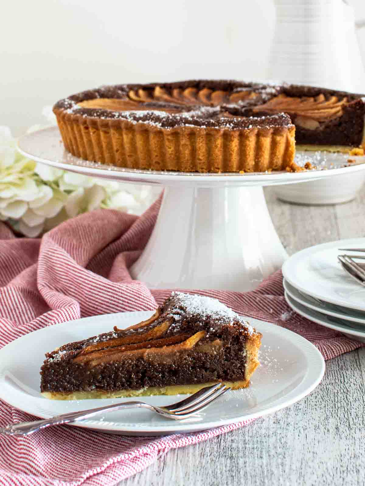 Chocolate almond pear tart on a white cake stand with a slice removed and plated in front.