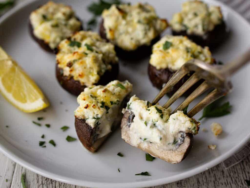 A plate of mushrooms caps stuffed with creamy mixture and one mushroom cut in half on a fork.