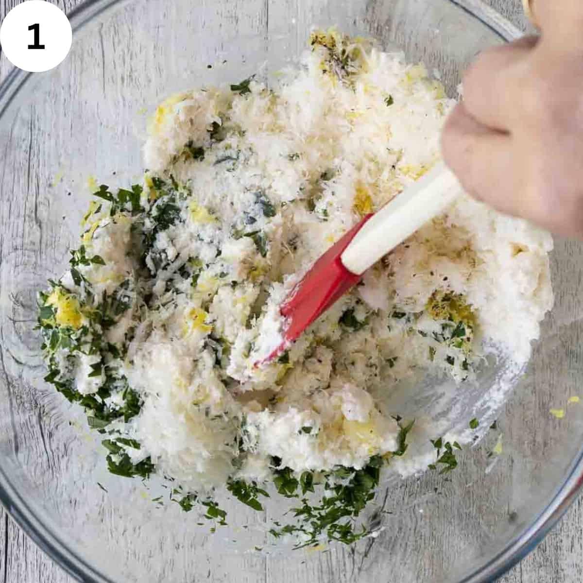 A spatula stirring ricotta, parmesan cheese and parsley in a glass bowl.