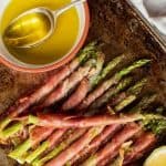 Prosciutto wrapped asparagus with lemon cheeks and olive oil in a red and white dish viewed from above.