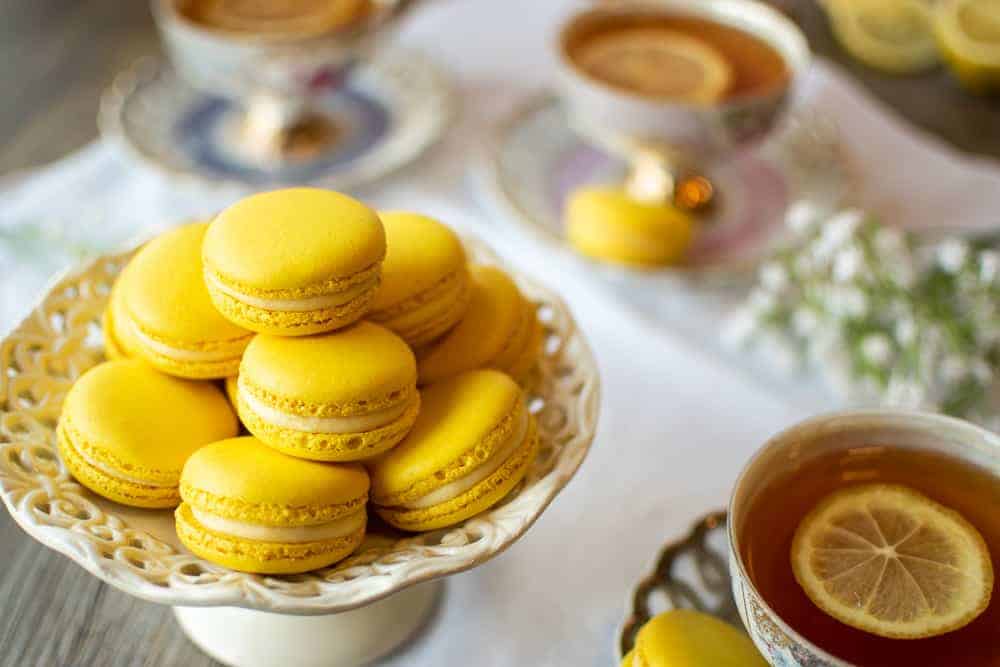 Lemon Macarons are filled with a tangy lemon flavoured white chocolate ganache #lemonmacarons #lemon #macarons #macaron Lemon Macarons on white stand with three cups of lemon tea on a white cloth