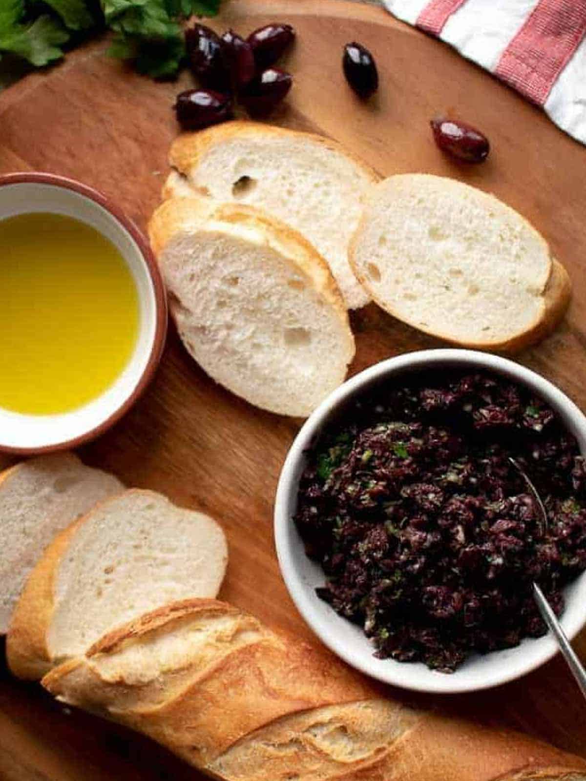 bowl of olivada with bread, oil and parsley on wooden board.