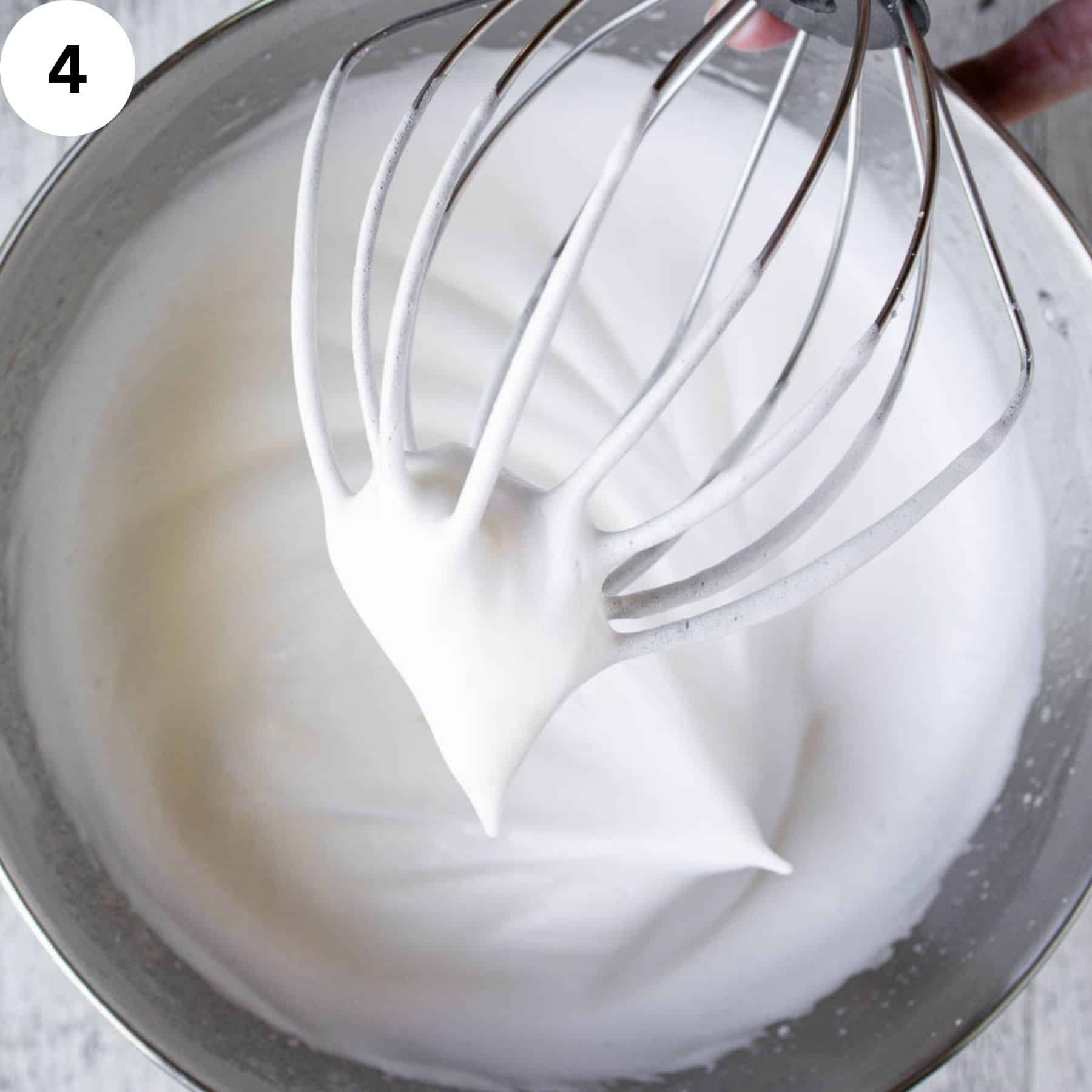 Whisk covered in stiffly beaten egg whites held above a bowl of beaten egg whites.
