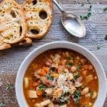 White bowl of slow cooker minestrone soup, two slices of toasted bread and spoon on wooden table