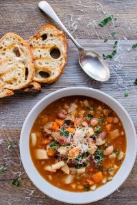 White bowl of slow cooker minestrone soup, two slices of toasted bread and spoon on wooden table