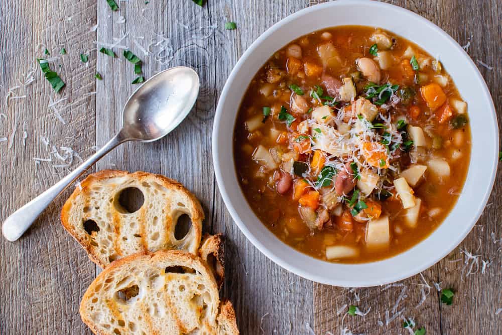 White bowl of slow cooker minestrone soup, two slices of toasted bread and spoon on wooden table