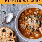 White bowl of slow cooker minestrone soup, two slices of toasted bread and spoon on wooden table