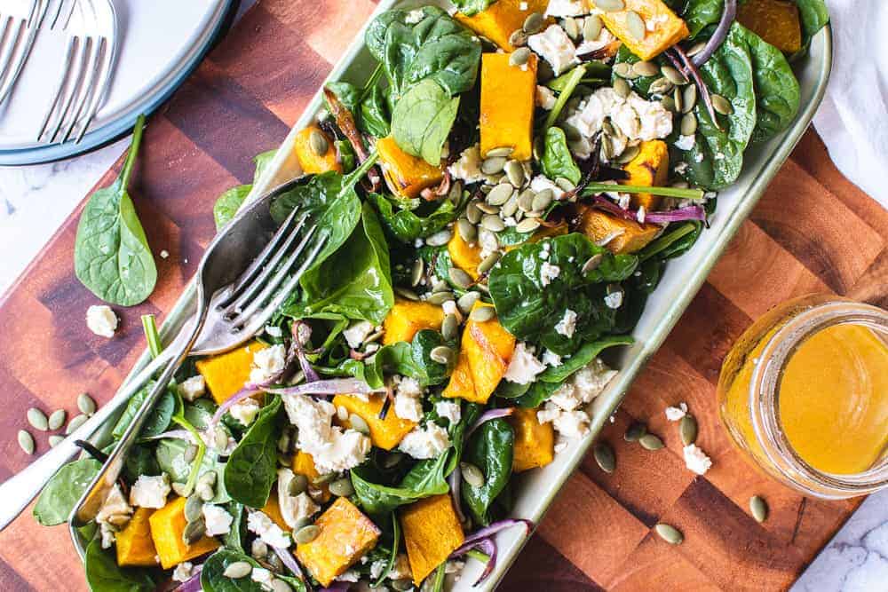 roast pumpkin salad on oblong plate on wooden board with salad dressing and plates and forks.