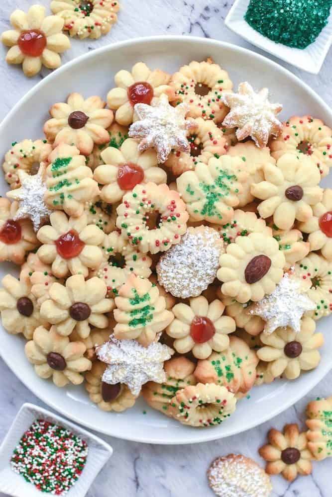 These buttery Spritz Cookies are delicious and not too sweet. The best thing about Spritz cookies is that they are easy and quick to make. #spritzcookies #cookies #butterycookies #christmascookies Decorated Italian butter cookies on a white plate with two small bowls of cake sprinkles.