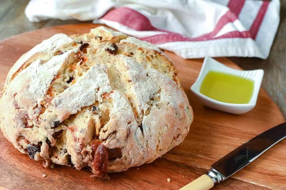 Damper bread on wooden board with knife, white dish of olive oil, dish clot in the background.