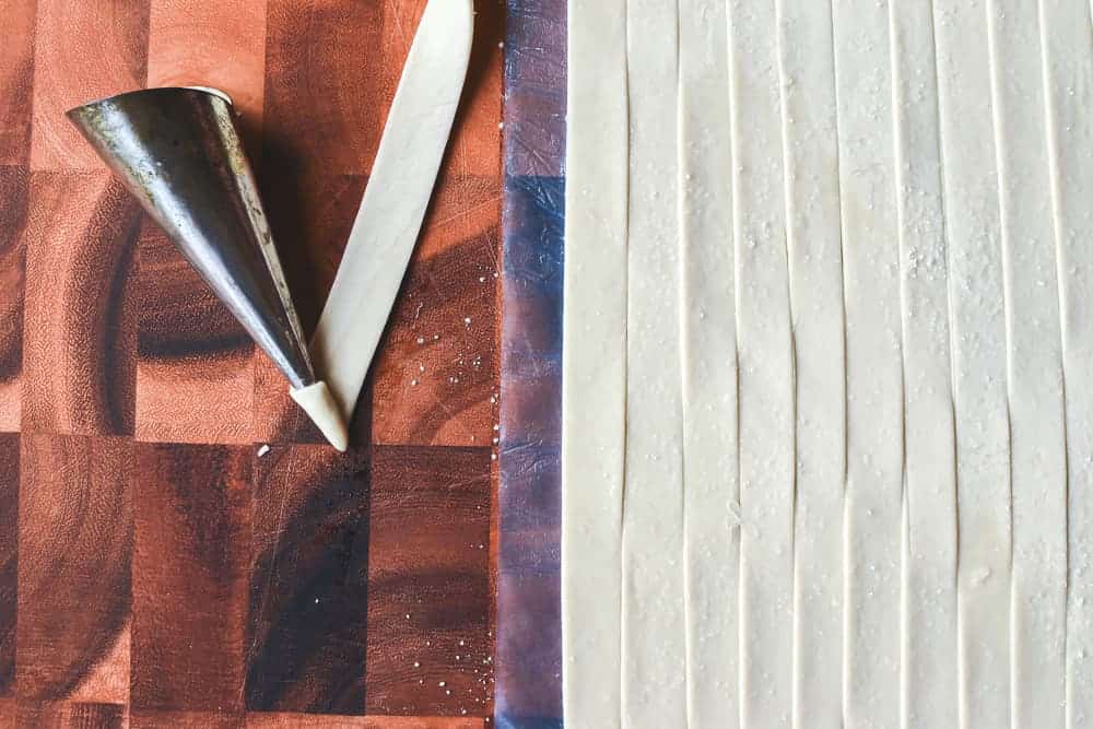 Sheet of raw puff pastry cut into strips with one strip being wound around a mould viewed from above.