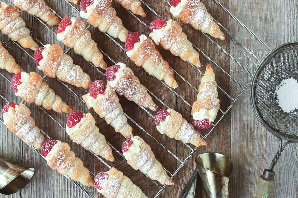 Mini cream horns filled with cream and topped with raspberry on a wire rack viewed from above.