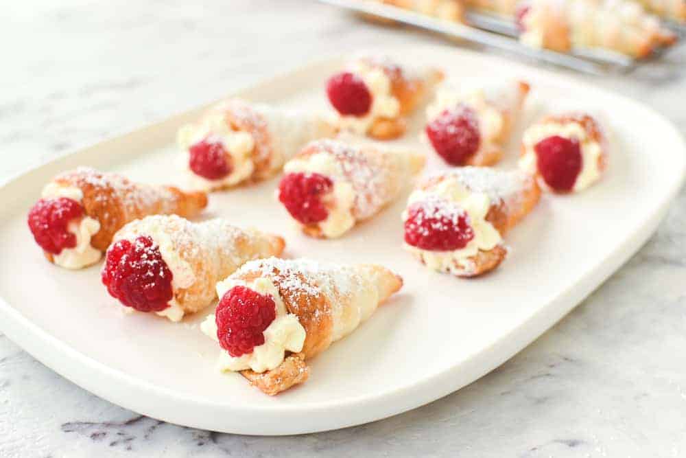 Puff pastry cones filled with cream and topped with raspberry on an oblong white plate viewed from an angle.