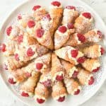 mini cream horns filled with cream and topped with raspberry arranged in a circular pattern on a round white plate viewed from above