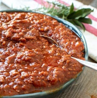 blue bowl overflowing with ragu pasta sauce, spoon in the sauce and herbs in the background