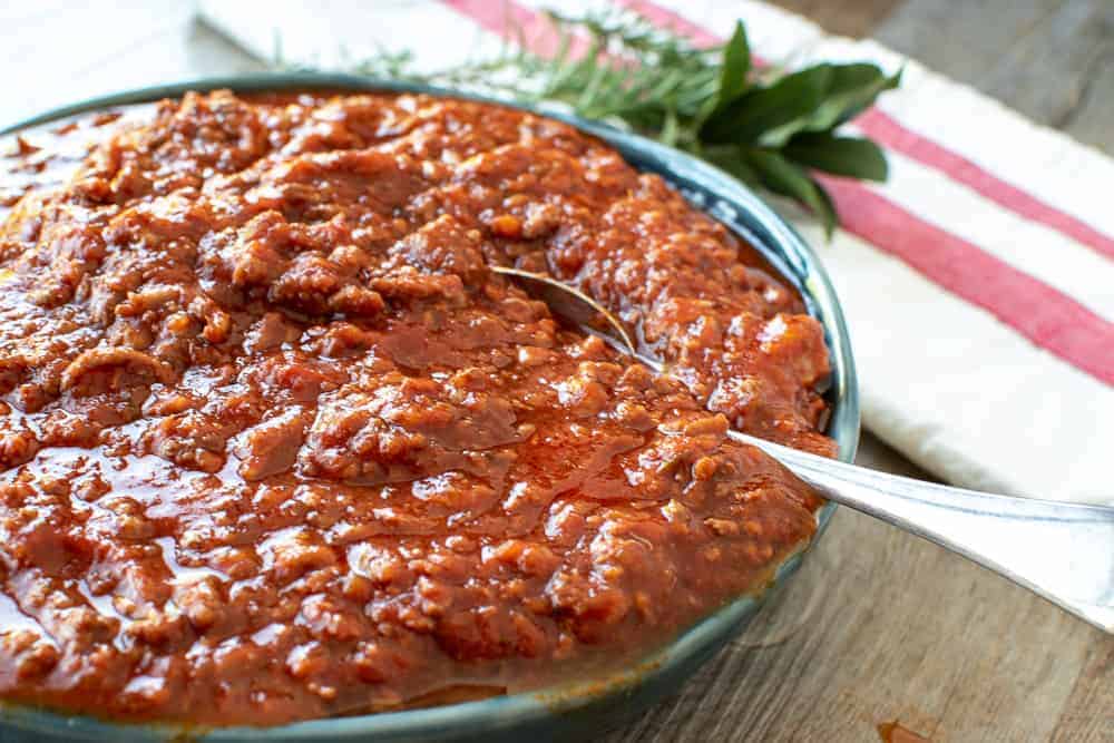 blue bowl overflowing with bolognese sauce, spoon in the sauce and herbs in the background.