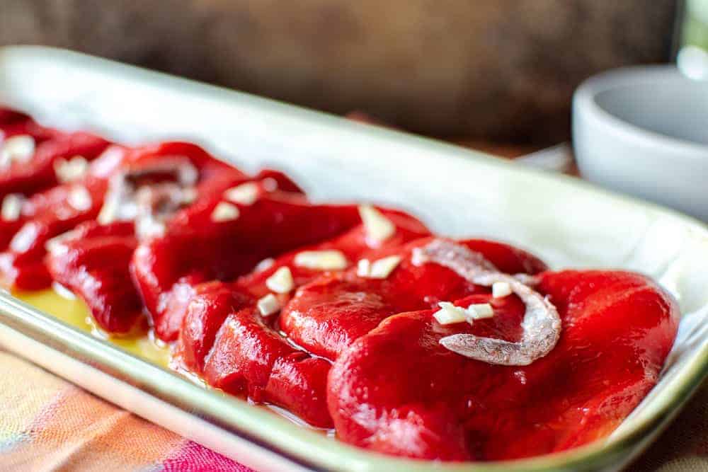 Roasted Red Peppers are easy to make at home. This recipe shows you how. #roastedredpeppers #recipe #peppers #italian #roasted Roasted Red Peppers on cream colored platter with a drizzle of oil, anchovies and a chopped garlic.