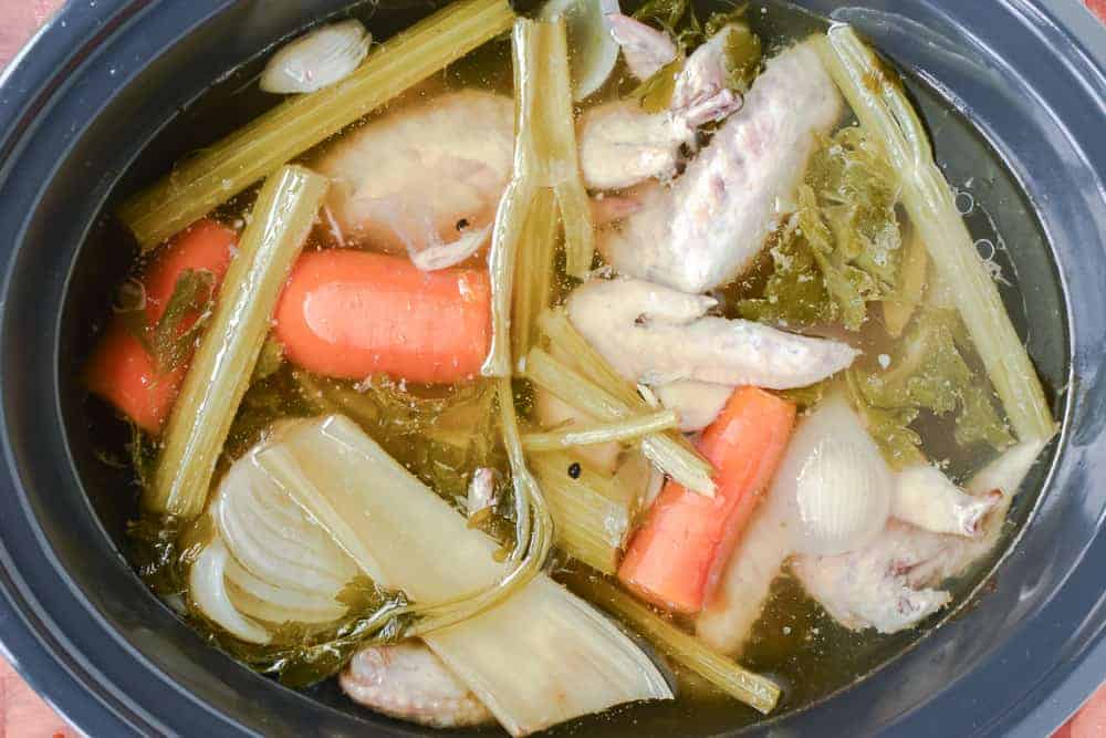 low cooker filled with chicken stock showing cooked chicken wings, carrots, celery, onions, parsley and garlic viewed from above