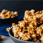 Homemade caramel popcorn in a grey bowl on blue cloth. smaller black bowl of caramel popcorn in background caramel popcorn and nuts on wooden table. close up