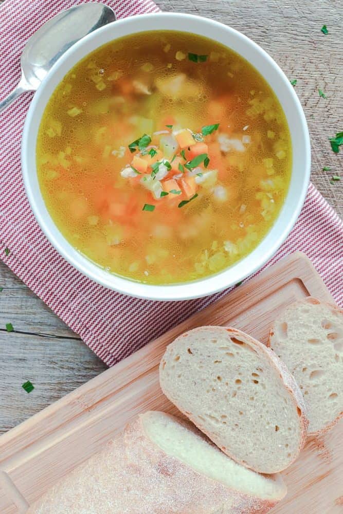 Chicken and vegetable soup in white bowl on red and white striped cloth, bread on wooden board viewed from above