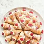 image with text. text reads "homemade cream horns. image is cream horns filled with cream and topped with raspberry arranged in a circular pattern on a round white plate viewed from above
